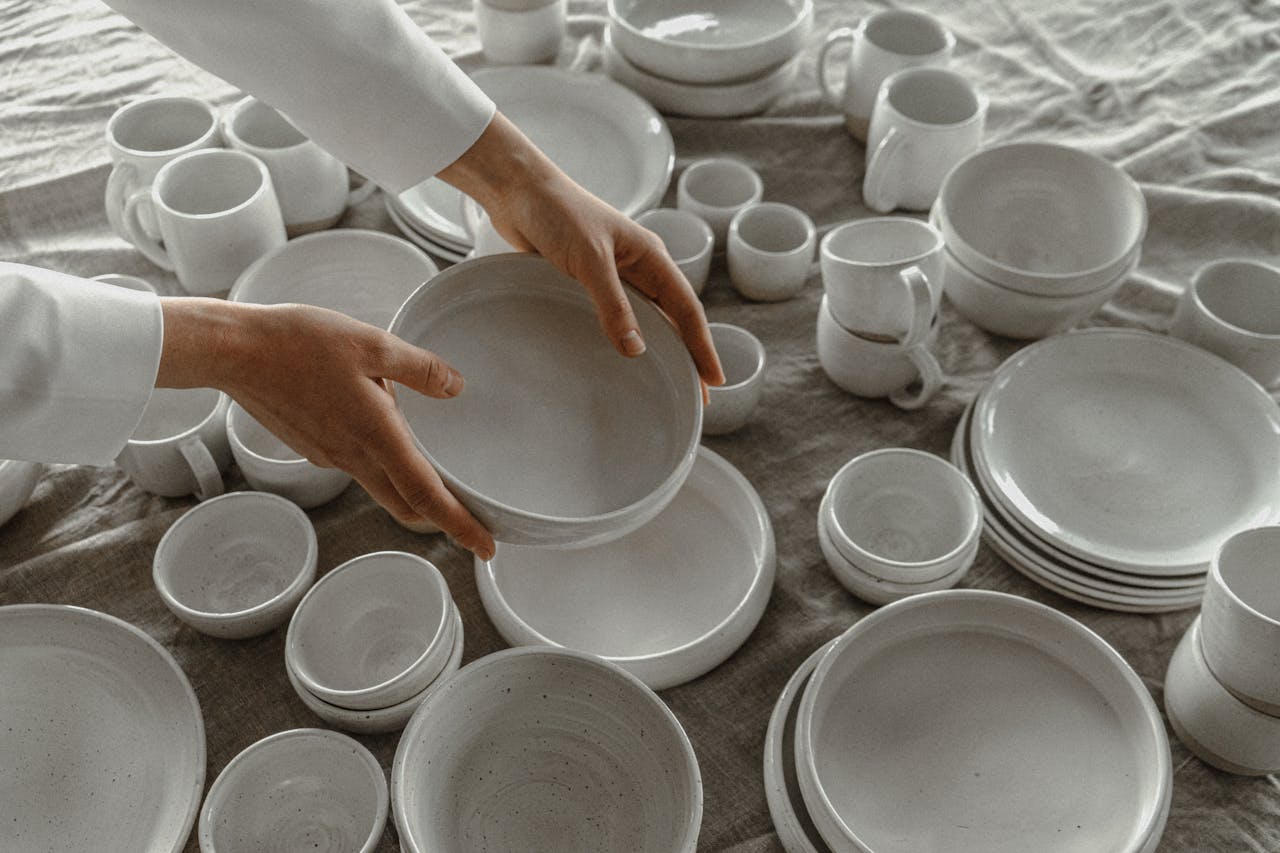 A close-up of hands arranging white ceramic dishware, showcasing elegant design on a textured tablecloth.